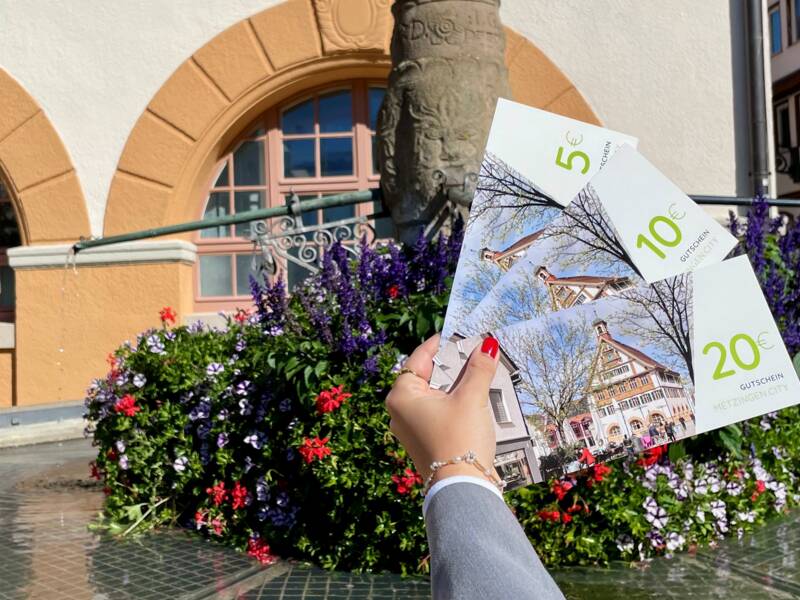 Eine Person hält drei Gutscheine über einem Blumenbeet vor einem Brunnen, im Hintergrund sind Gebäude mit markanten Rundbogenfenstern zu sehen.