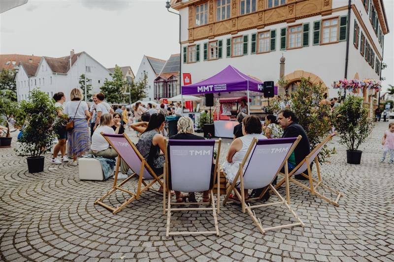 Eine Gruppe von Menschen sitzt auf einem gepflasterten Platz in Liegestühlen vor einem lilafarbenen Zelt mit der Aufschrift "MMT", umgeben von historischen Gebäuden und Passanten.